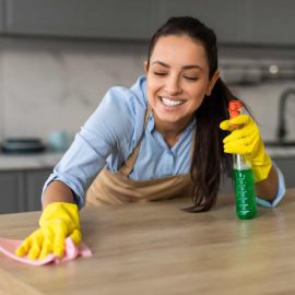 Cheerful woman in yellow gloves and an apron cleans kitchen counter with pink cloth and green spray bottle, smiling as she works, closeup